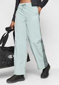 Šviesiai mėlyni sportiniai džinsai su raišteliu juosmens, šoninėmis baltomis juostelėmis ir mažuoju logotipu ant šlaunies; dėvimi su juodais sportiniais batais ir krepšiu.