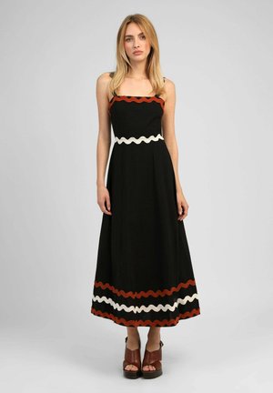 Femme debout portant une robe maxi noire sans manches avec des bordures ondulées rouges et blanches au niveau de la poitrine, de la taille et de l'ourlet, associée à des sandales compensées marron.