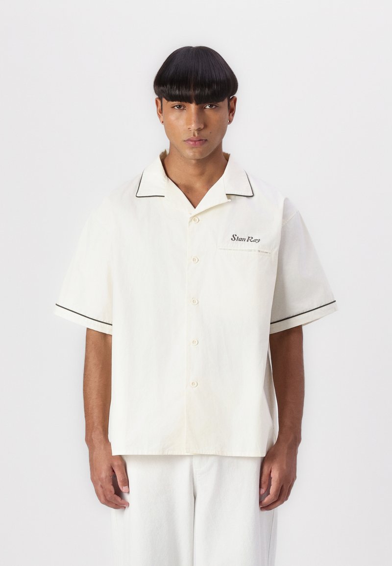 Short-sleeve, cream-colored shirt with contrasting black piping, button front, and a patch pocket. Smooth texture, minimal design.
