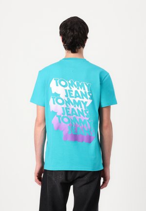 Turskizna pamučna majica s odvažnim, ponovljenim tekstom "TOMMY JEANS" u bijeloj i ljubičastoj gradijentu na leđima. Standardni kroj s kratkim rukavima.