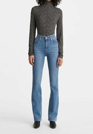 Femme portant un jean droit bleu clair taille haute, un col roulé noir ajusté à manches longues avec motif floral, et des chaussures noires à bout pointu, debout sur un fond blanc.