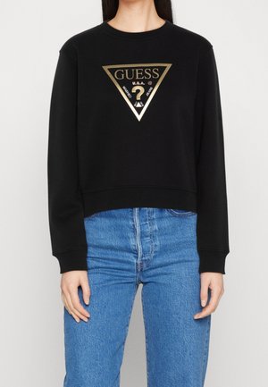 Frau trägt einen schwarzen Langarm-Sweatshirt mit einem goldenen dreieckigen Guess-Logo und hoch taillierte blaue Jeans.