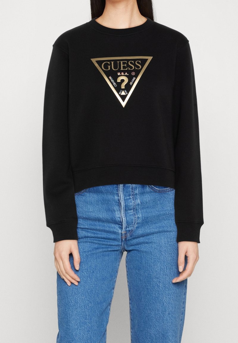 Femme portant un sweat-shirt noir à manches longues avec un logo triangulaire doré Guess et un jean bleu taille haute.