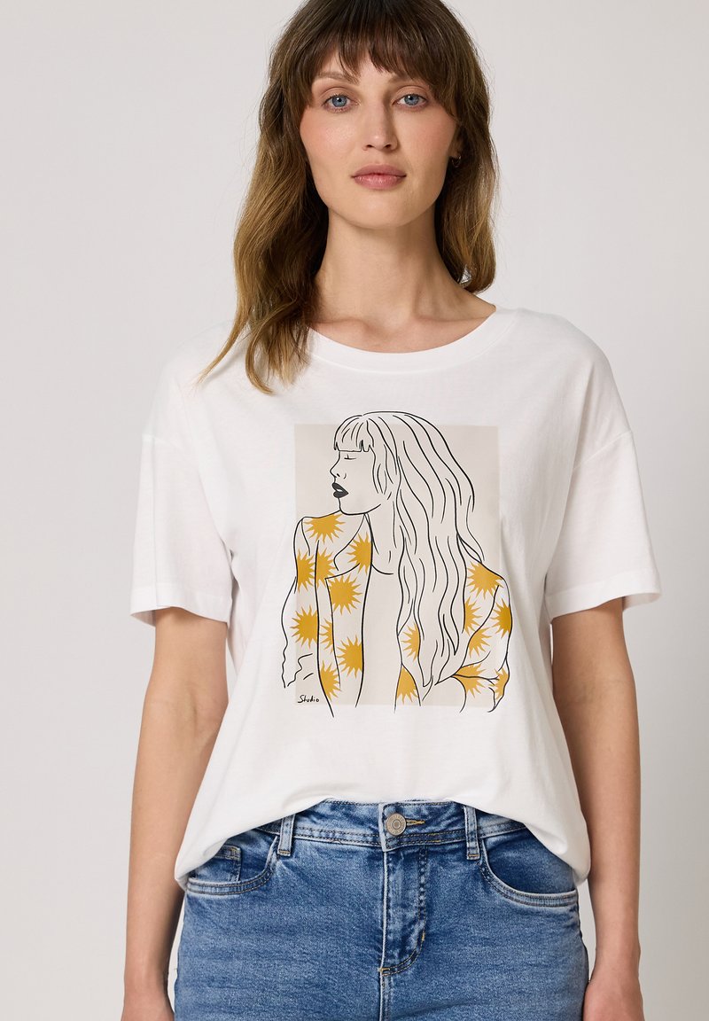 Femme portant un t-shirt blanc avec un portrait en ligne d'une femme aux longs cheveux et une veste jaune à motif floral, associée à un jean bleu.