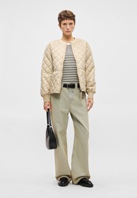 Ραφινάτη μπεζ bomber ζακέτα με καπιτονέ, ριγέ πουκάμισο, φαρδιά ανοιχτόχρωμα τζιν και μαύρη τσάντα, φορεμένα με μαύρα παπούτσια.