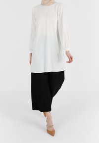 Weißes langärmliges Oberteil mit plissiertem Rückendetail, kombiniert mit lockeren schwarzen Hosen und beigen spitz zulaufenden High Heels. Einheitliche Textur ohne Muster.