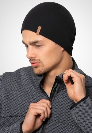Czarna dzianinowa czapka beanie z małą skórzaną naszywką z boku, noszona przez osobę w teksturowanej szarej kurtce z zamkiem błyskawicznym i wysokim kołnierzem.