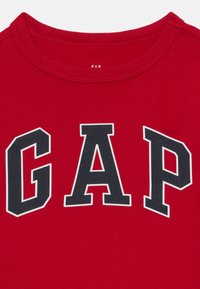 Raudonas medvilnės marškinėliai su dideliu juodu "GAP" logotipu su baltu apvadu. Klasikinis apvalus kaklas ir lygus tekstūros paviršius.