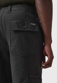 Pantaloni cargo neri realizzati in tessuto testurizzato, con una tasca laterale dotata di chiusura a bottone e un'etichetta "ALLSAINTS" sul retro.