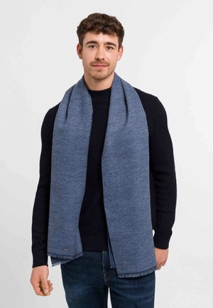 Man wearing a textured blue scarf over a black sweater and blue jeans, standing against a plain white background.