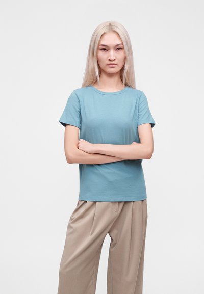Jeune personne aux longs cheveux blonds se tient les bras croisés, portant un t-shirt bleu et un pantalon beige sur un fond blanc uni.