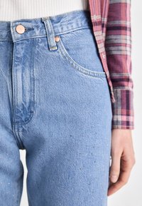 Svijetloplave traperice sa ravnim nogavicama, ukrašene bakrenim dugmadi i dekorativnim perlicama sprijeda. Vidljiva suptilna tekstura.