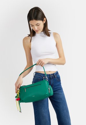 Giovane donna con top bianco senza maniche e jeans blu che tiene una borsa verde con portachiavi con nappine colorati su sfondo semplice.