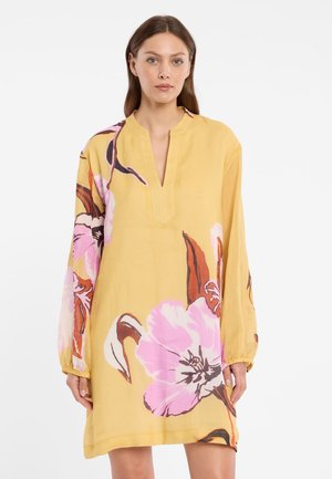 Femme portant une robe ample jaune avec de grandes impressions florales roses et marron, manches longues et encolure en V.