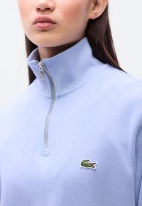 Ανοιχτό μπλε φούτερ με φερμουάρ και ψηλό γιακά, με ριμπ υφή, που διαθέτει ένα μικρό πράσινο κεντητό λογότυπο κροκόδειλου στο αριστερό στήθος.