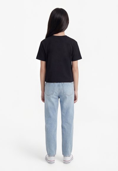 Fille debout de dos portant un t-shirt noir, un jean bleu clair et des baskets blanches sur un fond blanc uni.