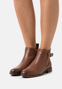 Bottines en cuir marron avec un bout arrondi, fermeture éclair sur le côté et une accent de boucle à la cheville. Talon bas empilé et texture lisse.