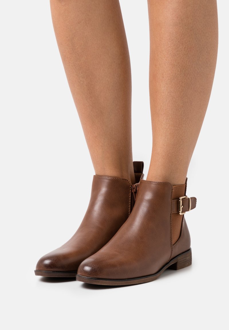 Bottines en cuir marron avec un bout arrondi, fermeture éclair sur le côté et une accent de boucle à la cheville. Talon bas empilé et texture lisse.