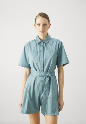 Pantalon bleu à manches courtes avec un col, deux poches avant, des boutons et un détail de ceinture nouée à la taille. Fabriqué en tissu léger.