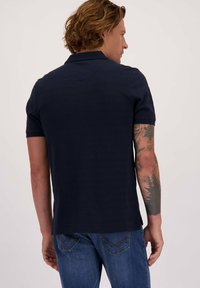 Man met golvend haar, gekleed in een marine blauwe polo en blauwe spijkerbroek, met een gedetailleerde bos thema tatoeage op zijn rechter onderarm.