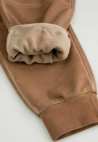 Złożone brązowe spodnie dresowe z widoczną teksturowaną beżową podszewką i ściągaczem na kostce na jasnym tle.