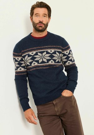 Hombre con barba que lleva un suéter azul marino con patrones blancos de copos de nieve y pantalones marrones, de pie contra una pared lisa y clara.