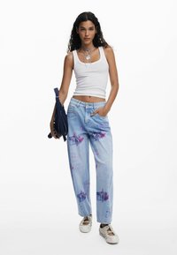 Jeans in denim azzurro chiaro con effetto sbiadito e stampa grafica viola, abbinati a una canotta bianca. Gli accessori includono una borsa scura.