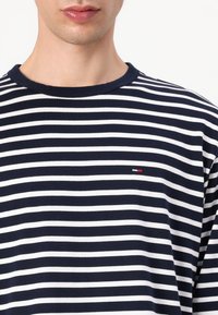Marineblå og hvite stripete langermet t-skjorte med rund hals, med en liten logo i rødt, hvitt og blått på venstre side.