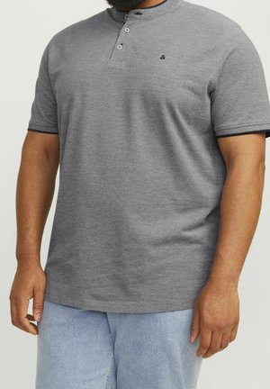 Mann trägt ein graues Kurzarm-Henley-Shirt mit schwarzem Besatz an den Ärmeln und hellblauen Jeans, steht vor einem einfarbigen Hintergrund.