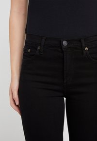 Jeans de mezclilla negra con un diseño ajustado. Estilo de cinco bolsillos con herrajes en tono dorado y cierre de botón en la cintura. Textura suave.