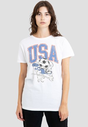 Junge Frau trägt ein weißes T-Shirt mit großen blauen und roten USA-Buchstaben und einer Comicfigur, die vorne Fußball spielt, vor einem einfarbigen Hintergrund.