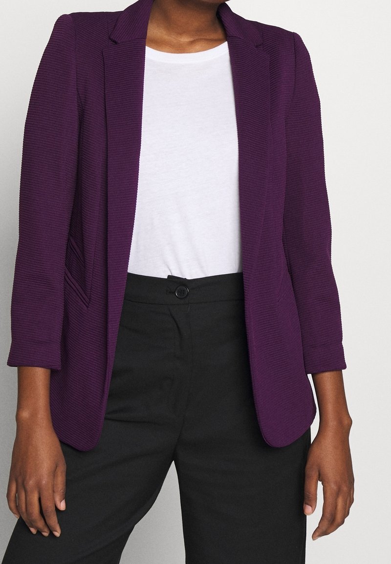 Femme en blazer violet texturé, chemise blanche à col rond et pantalon noir taille haute, debout les bras détendus le long du corps.
