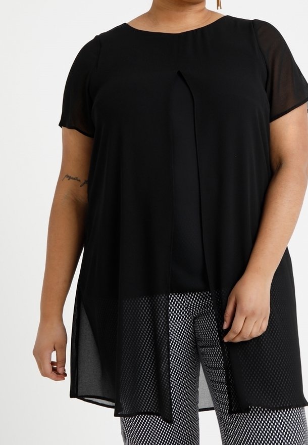 Femme portant une tunique noire transparente à manches courtes par-dessus un top noir et un pantalon noir et blanc à motifs sur un fond uni.