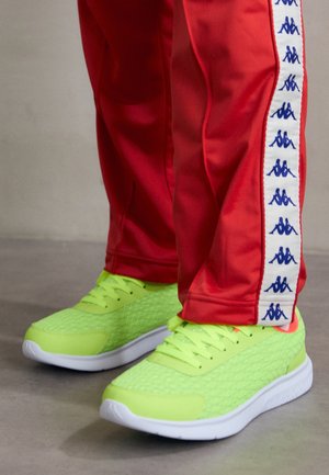Baskets jaune vif portées avec un pantalon de survêtement rouge présentant une bande blanche avec des logos bleus sur le côté.