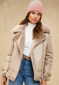 Beige shaggy jakker laiaga kraega ja esi lukuga. Seljas valge soonikkoes särk ja sinised teksad, lisatud roosa mütsiga.
