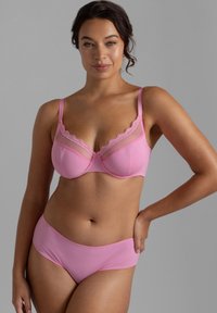 Γυναίκα με σκουρόχρωμα μαλλιά που φοράει ένα ροζ σετ εσώρουχα με δαντέλα στα κύπελλα του στηθόδεσμου, στέκεται με το ένα χέρι στη μέση της μπροστά σε γκρι φόντο.