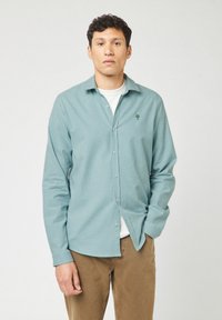 Lichtblauw geruit overhemd met lange mouwen en een knoopsluiting kraag, met een groen boomlogo op de borst, gecombineerd met een wit T-shirt eronder.