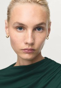 Mladá žena s blond vlasy staženými dozadu, s lehkými pihami, zlatými kruhovými náušnicemi, oblečená v tmavě zeleném topu, hledící přímo vpřed s neutrálním výrazem.