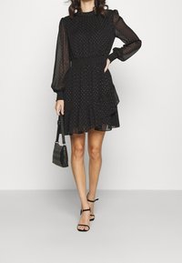 Vestido negro de Manga Larga con cintura fruncida y falda de volantes en capas. Presenta sutiles lunares y un cuello alto. Combinado con un bolso negro.