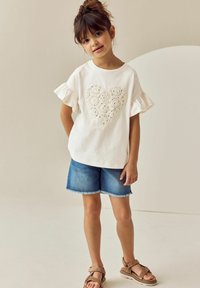 Weißes Baumwoll-T-Shirt mit Rüschensleeves und einem dekorativen Häkelherz, kombiniert mit blauen Denim-Shorts und braunen Riemchensandalen.