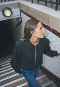 Zwarte jas met rits en een gestructureerde kraag, gecombineerd met een blauwe spijkerbroek, gedragen door een persoon die een trap afloopt met witte betegelde muren.