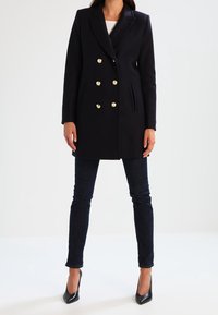 Dubbelt knäppt svart ullrock med guldfärgade knappar, ficklappar, sidofickor och en skräddarsydd passform, i kombination med mörka jeans och svarta klackar.