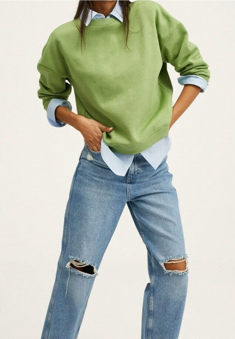 Femme portant un pull vert clair par-dessus une chemise bleu clair aux manches retroussées et un jean bleu déchiré, mains posées sur les hanches.