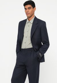 Σκούρο σκούρομπλε σακάκι με πέτα, σε συνδυασμό με ανοιχτό γκρι πουκάμισο με κουμπιά. Η εμφάνιση χαρακτηρίζεται από λεπτές υφές και εφαρμοστή κοπή.