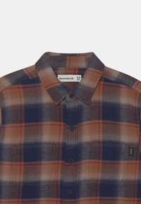 Rutigt skjorta med knappar i brunt, marinblått och orange. Har krage, två bröstfickor och svarta knappar. Tillverkad av mjukt tyg.