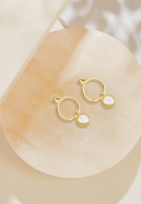 Gyldna hoopörhängen har en rund design med en klar oval stenpendant som sitter på en slät neutral yta.