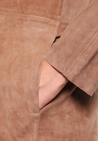 Brun mockaskinnjacka med detalj av en hand i fickan, som framhäver den mjuka texturen och synliga sömmar längs med sömmarna.