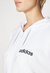 Fehér, cipzáros kapucnis pulóver, zsinórokkal, elöl fekete "adidas" logóval. Simogató anyag, laza szabás.