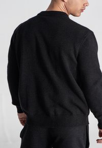 Mann in einem schlichten schwarzen Langarmpullover und passenden schwarzen Hosen, der mit dem Rücken zur Kamera steht und den Kopf seitlich dreht.