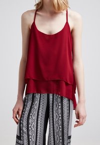 Burgunderfarbene Satin-Camisole mit dünnen Trägern, schichtweise gestaltet, hinten etwas länger, getragen mit schwarz-weißen gemusterten weit geschnittenen Hosen.